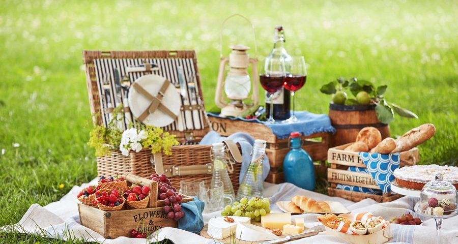 Wicker picnic hamper with assorted fresh food, infused water and wine on a rug spread out on the green grass in a park
